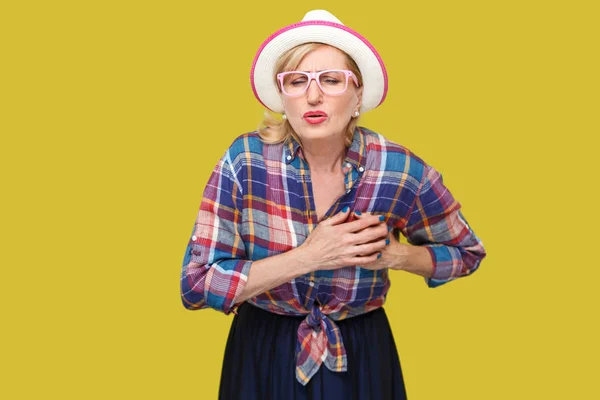成熟的女人在休闲风格与帽子和眼镜抱着她的乳房 而感觉疼痛的心脏在黄色背景 — 图库照片