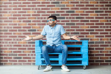 Rahat tarzı ve gözlük mavi ahşap palet üzerinde oturan ve kahverengi tuğla duvar arka plan üzerinde boşanmış kolları ile uzağa bakarak karışık yakışıklı genç sakallı adam tam uzunlukta 