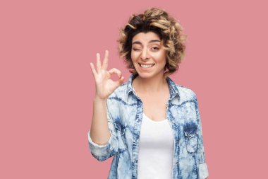 funny young woman with curly hairstyle in casual blue shirt showing Ok sign gesture hand and winking with toothy smile on pink background