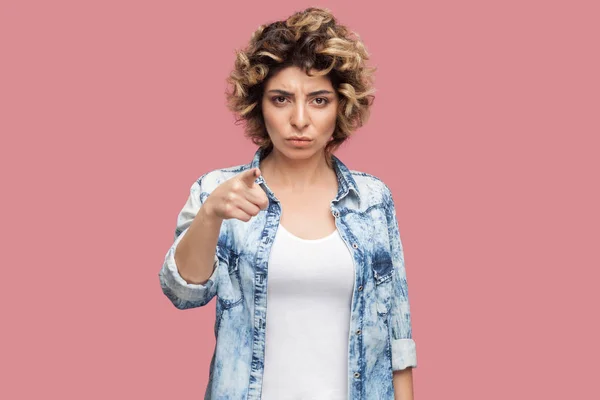 serious young woman with curly hairstyle in casual blue shirt looking and pointing at camera with serious face on pink background