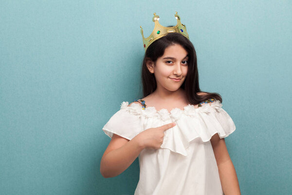 arrogance beautiful brunette girl wear in white dress and golden diadem crown standing and pointing finger to herself on blue background