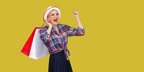 白い帽子をかぶった現代のおばあさんとショッピングバッグを抱えた格子柄のシャツに 黄色い背景に微笑みながら歯ながら勝ち抜く — ストック写真