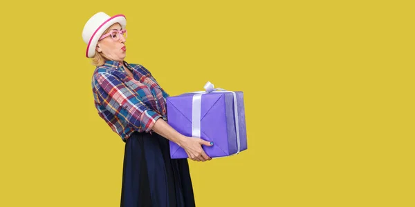 Side view of cheerful wondering modern grandmother in white hat and checkered shirt trying to hold giant big heavy gift box on yellow background