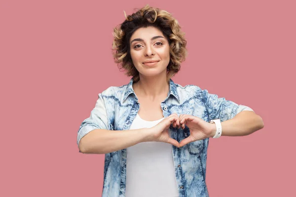 happy young woman with curly hairstyle in casual blue shirt showing heart hands shape gesture and looking at camera while smiling on pink background