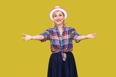 mutlu huzurlu modern şık olgun kadın rahat tarzı şapka ile ayakta kolları ile ve sarı arka plan üzerinde dişli gülümseme ile kamera bakıyor