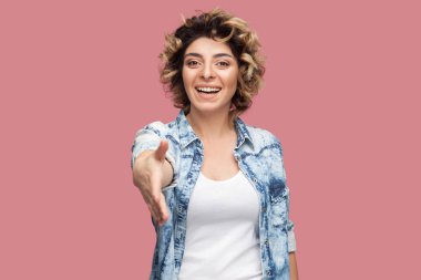 rahat mavi gömlek dişli gülümseyerek kıvırcık saç modeli ile mutlu genç kadın gülümseyerek ve pembe arka plan üzerinde tebrik ve el sıkışma elini veren