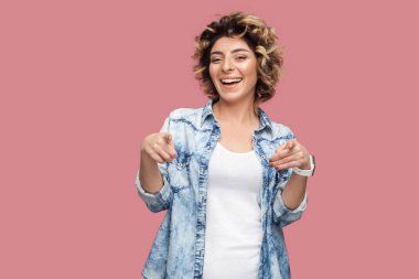 rahat mavi gömlek kıvırcık saç modeli ile mutlu komik genç kadın görünümlü ve pembe arka plan üzerinde dişli gülümseme ile kamera işaret