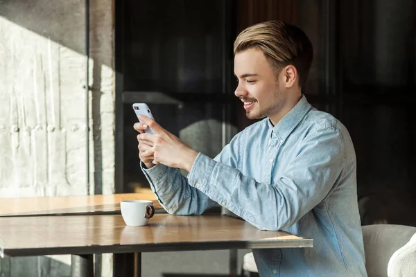 カフェに座って 歯の笑顔でスマートフォンでメッセージを読んで青いデニムシャツを着たハンサムな幸せな若者の側面図 — ストック写真