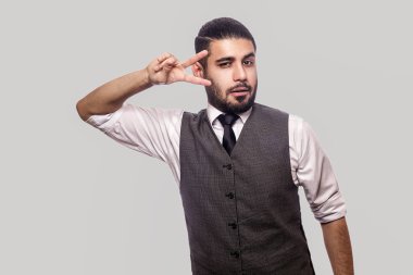 beyaz gömlek ve yelek zafer işareti gösteren ve gri arka planda kamera bakarak komik yakışıklı sakallı esmer adam.