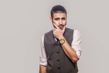 beyaz gömlek ve yelek yüze dokunmadan karışık yakışıklı sakallı esmer adam ve gri arka planda üzgün yüzü ile kamera bakıyor.