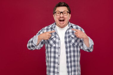 rahat damalı gömlek ve gözlük yakışıklı orta yaşlı adam kamera bakarak ve koyu kırmızı arka plan üzerinde kendini işaret şaşırttı