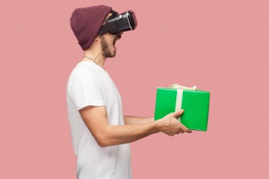 Beyaz gömlek ve rahat şapka vr kulaklık giyen şok sakallı genç hipster yan görünümü ve pembe arka plan üzerinde açık ağız ile yeşil mevcut kutu tutan