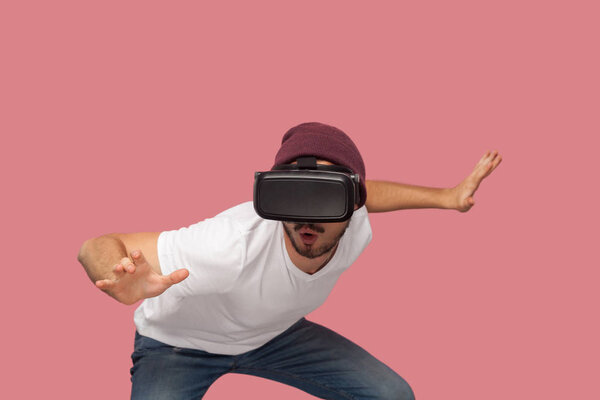 shocked bearded young man in white shirt and casual hat wearing vr headset playing video game with raised arms on pink background