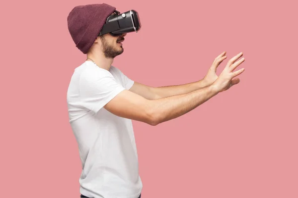 Side view of shocked bearded young man in white shirt and casual hat wearing vr headset playing video game on pink background