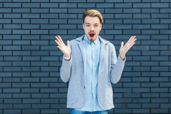 shocked handsome young blond man in casual style standing with raised arms and looking at camera with unbelievable surprised face on brick wall background