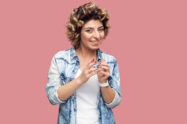 rahat mavi gömlek ayakta kıvırcık saç modeli ile komik kurnaz genç kadın ayakta ve pembe arka plan üzerinde dişli gülümseme ve eğlenceli yüz ile kamera bakıyor