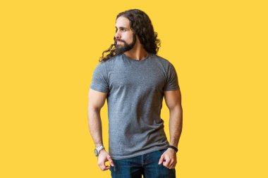 gri t-shirt ayakta uzun kıvırcık saçlı acımasız yakışıklı sakallı genç adam model ayakta ve sarı arka plan üzerinde ciddi gurur yüzü ile uzağa bakıyor