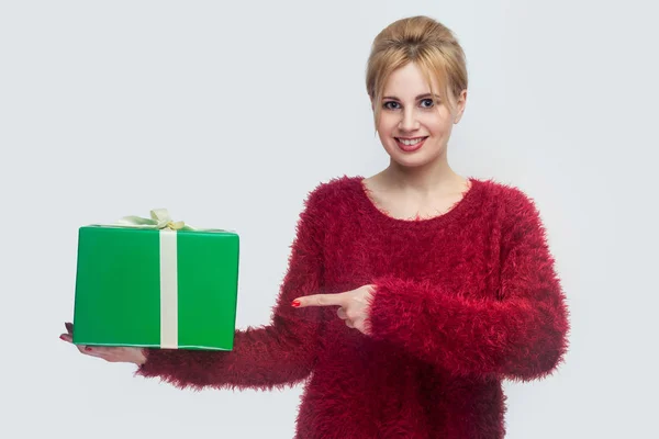 穿着红色毛衣的快乐年轻女子指着手中的绿色礼品盒 看着灰色背景上的相机 — 图库照片