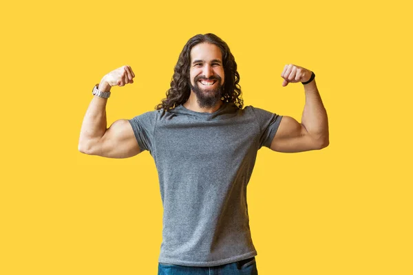 happy strong bearded young bodybuilder man with long curly hair in grey t-shirt showing muscular arms and looking at camera with toothy smile on yellow background