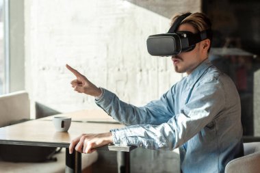 mor denim gömlekli sürpriz sakallı genç yetişkin adam vr kulaklık giyen ve simülatörvideo izlerken parmak kenara işaret ederken ve kafede otururken , eğlence yaşam tarzı.
