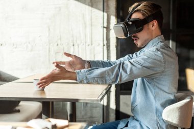 vr kulaklık giyen ve kafede kolları ve açık ağız ile otururken simülatör video izlerken mavi denim gömlek sürpriz sakallı adam yan görünümü , eğlence yaşam tarzı.