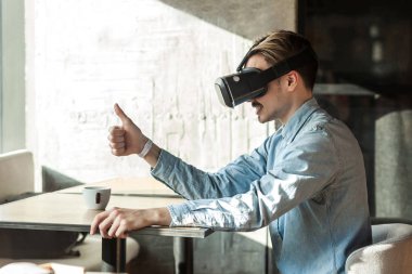 vr kulaklık giyen ve kafede otururken başparmak gösteren mavi denim gömlek mutlu adam yan görünümü , eğlence yaşam tarzı
