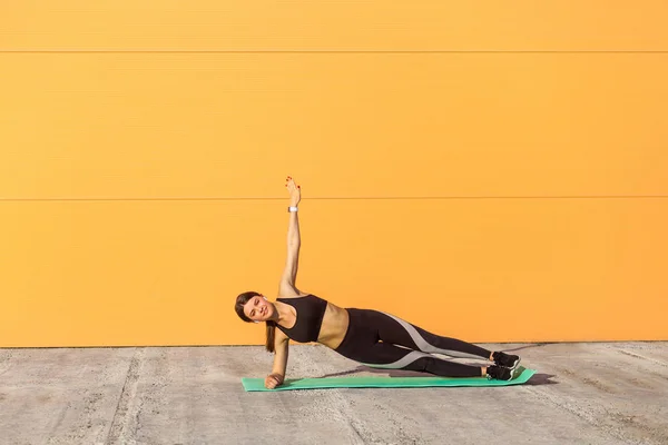 ヨガを練習しながら オレンジ色の壁の背景 スポーツ 健康的な概念に対して日光の下でバシスタサナ運動をしながら 若いスポーティな魅力的な女性が肘にサイドプランクポーズを立っています — ストック写真