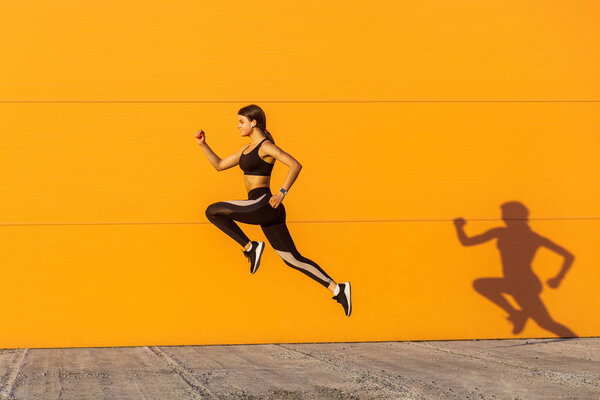 Young sporty beautiful woman with fit body jumping and running  in sunshine against orange wall background