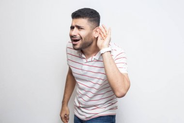 özenli yakışıklı sakallı genç adam çizgili t-shirt kulak üzerinde el ile ayakta ve beyaz arka plan üzerinde izole duymaya çalışıyor