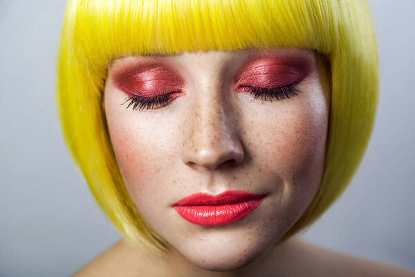 Beauty portrait of cute young woman with closed eyes and red makeup in yellow wig with serious on gray background 