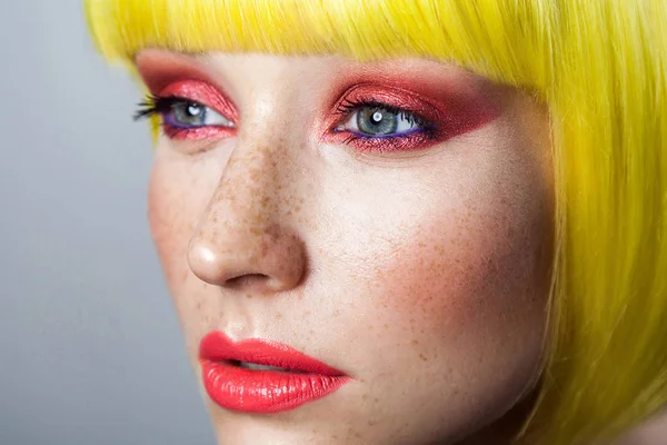 Beauty portrait of calm cute young female model with freckles and red makeup in yellow wig on gray background