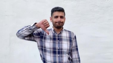 mavi damalı gömlek memnun yakışıklı sakallı adam kameraya bakıyor ve beyaz duvar arka planda başparmak aşağı gösteren