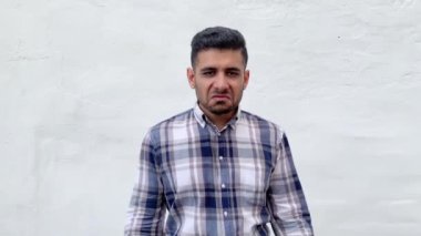 Mavi kareli gömlekli kızgın, yakışıklı, sakallı adam kameraya bakıyor ve beyaz duvar arkasında orta parmak işareti gösteriyor.