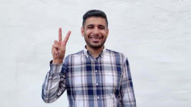 zafer jestel işareti gösteren mavi damalı gömlek mutlu yakışıklı sakallı adam ve beyaz duvar arka plan üzerinde dişli gülümseme ile bakıyor