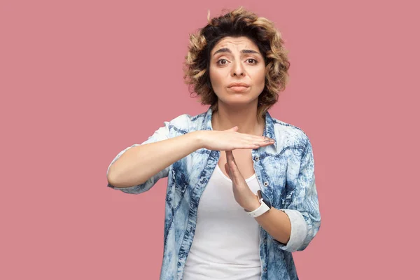 worry pleased young woman with curly hairstyle in casual blue shirt showing timeout gesture sign and looking at camera on pink background