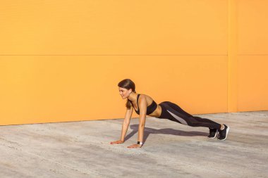 Turuncu duvar arka plan, spor ve sağlıklı bir konsepte karşı mükemmel plaka lı pozisyonda duran siyah spor giyimli kaslı sportif güçlü genç atletik kadının yan görünümü