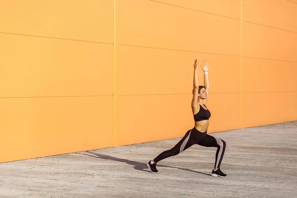 黒いスポウトを着てヨガを練習し オレンジ色の壁の背景 スポーツ 健康的な概念にビラバドラサナポーズをしながら 戦士の一つの運動に立っている若い魅力的な女性の側面図 — ストック写真
