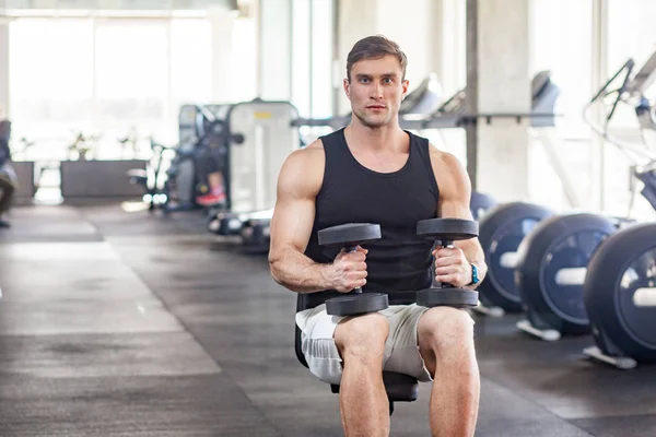 young adult muscular built handsome athlete sitting on weightlifting machine and holding two dumbbell on knees while working out in gym