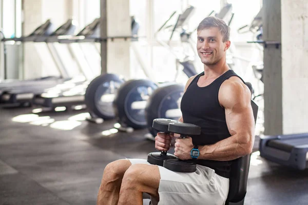 侧视图年轻的成人肌肉建设英俊的运动员坐在举重机上 并拿着两个哑铃跪 而在健身房锻炼 — 图库照片