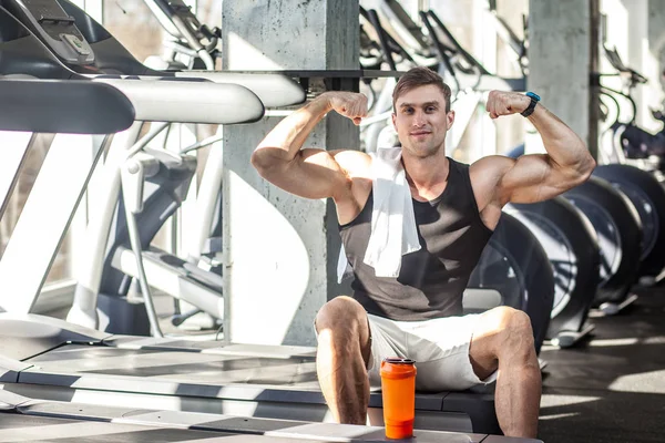 成功的运动男性男子看着相机 并显示肌肉凸起的手臂 而坐在跑步机锻炼在健身房 — 图库照片