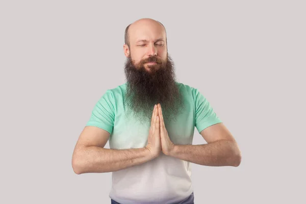 灰色の背景にヨガの瞑想をしながら 目を閉じて手のひらを握る薄緑色のTシャツで長いあごひげを持つ穏やかな真剣な中年ハゲ男 — ストック写真