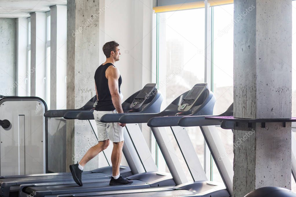 side view of young adult sport man walking on treadmill and looking to window while training at gym, healthy fitness concept