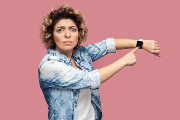 serious bossy young woman with curly hairstyle in casual blue shirt standing and pointing at her smart watch on pink background