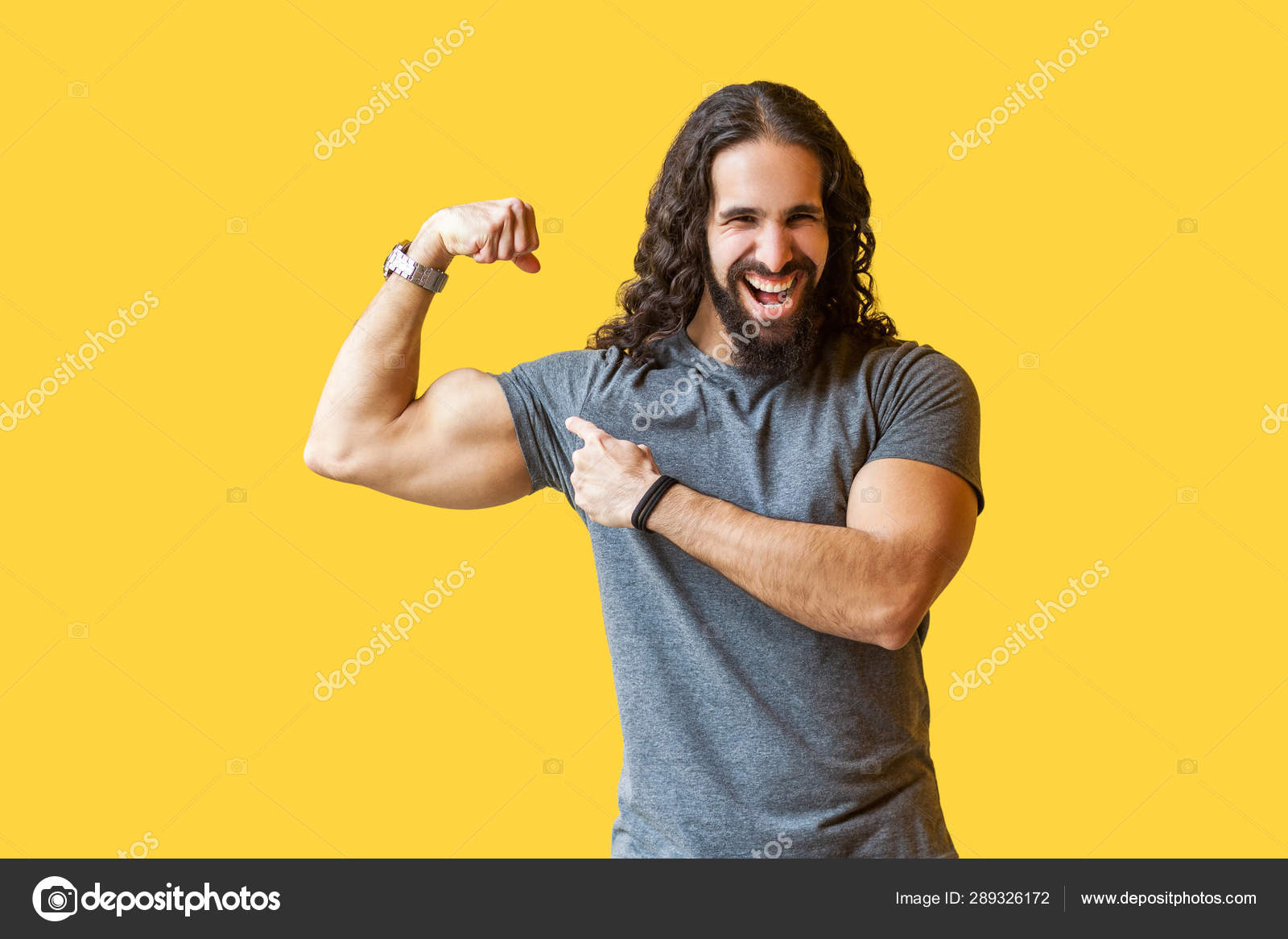 搞笑强胡子健美男子与灰色t恤长卷发指向他的二头肌看着相机与黄色背景的牙齿微笑 图库照片 C Khosrork