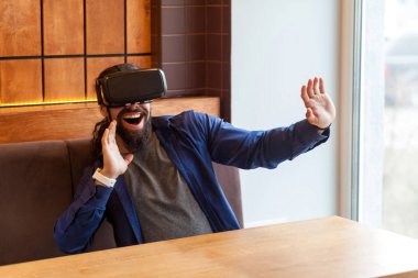 inanılmaz mutlu yüz ve yükseltilmiş kollar ile simülatörü video izlerken rahat tarzı kafede oturan ve VR kulaklık giyen mutlu inanılmaz sakallı genç yetişkin adam