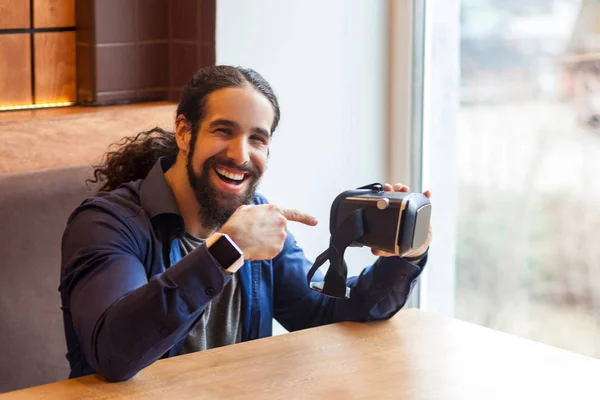 happy bearded young adult man in casual style sitting in cafe and pointing finger on vr headset while looking at camera with smile