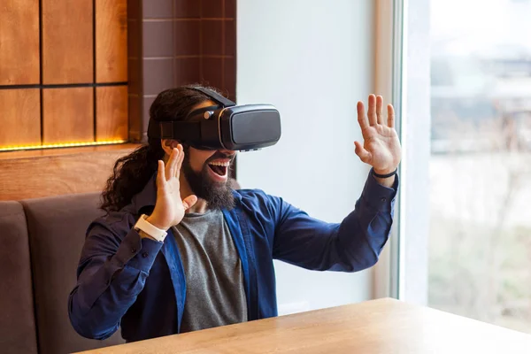 happy unbelievable bearded young adult man in casual style sitting in cafe and wearing vr headset while watching video on simulator with amazing happy face and raised arms