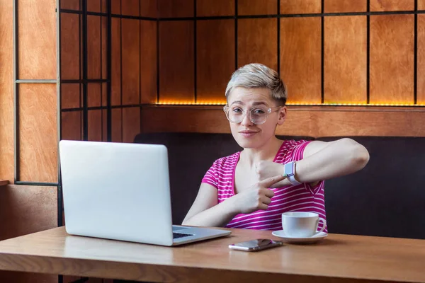 upset young bossy woman with blonde short hair in pink t-shirt and eyeglasses sitting with laptop in cafe and showing checking time gesture while pointing finger to wrist watch