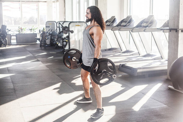 Вид сбоку на молодого взрослого культуриста с длинными вьющимися волосами, занимающегося спортом, стоя и готовясь к поднятию штанги, концепция здорового образа жизни
  