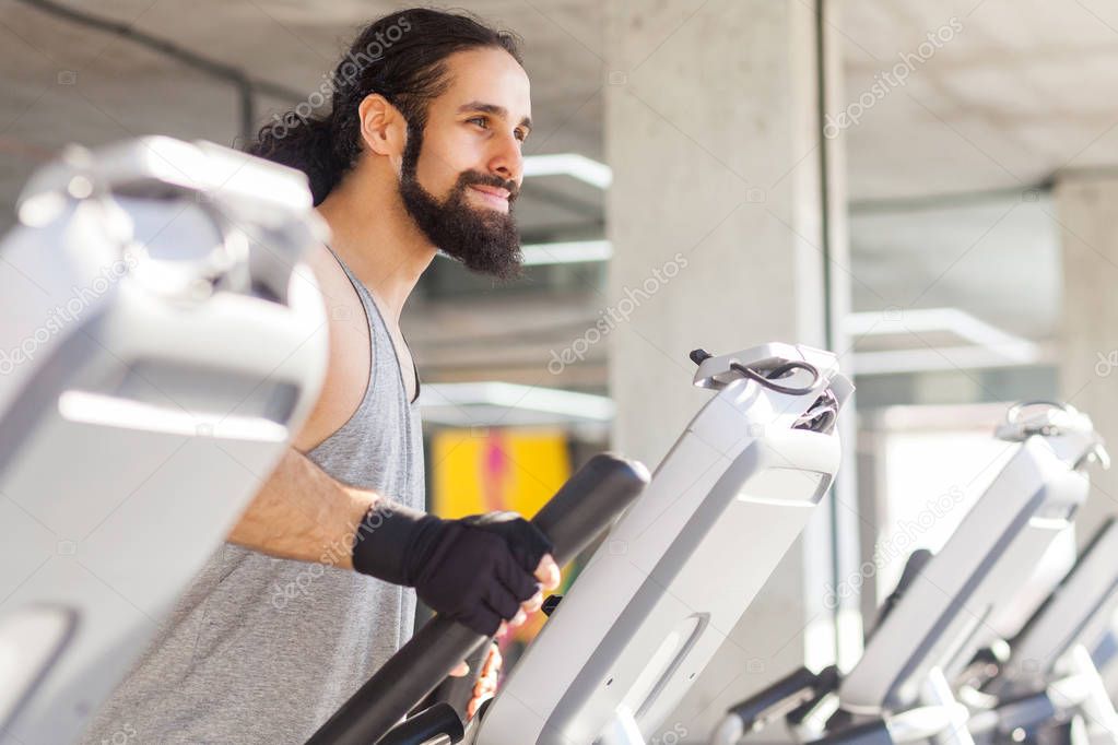 side view of young adult sportsman with long curly hair training on elliptical machine in gym, fitness concept